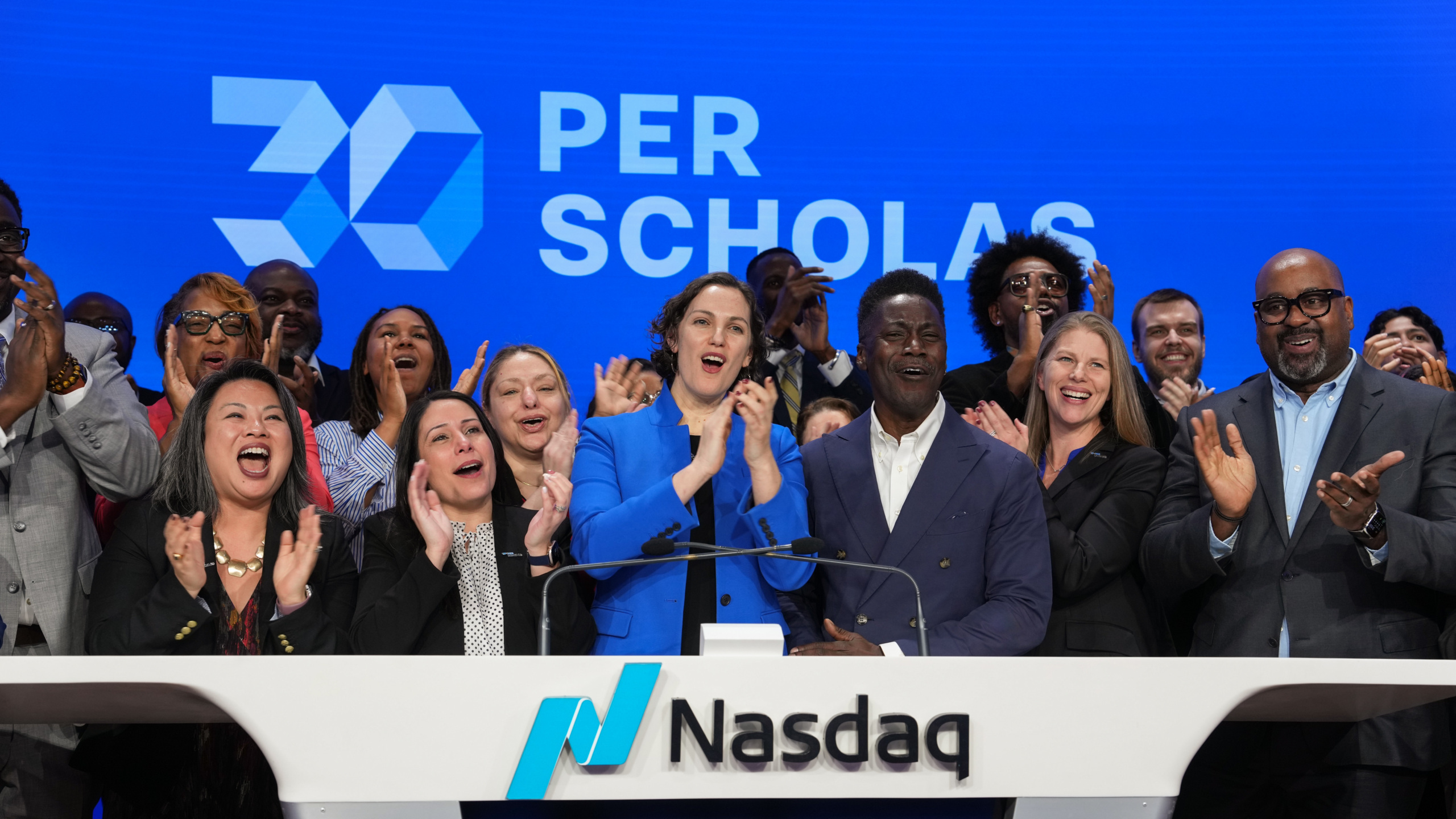 Group of adults cheering together onstage at the Nasdaq podium beneath a large "Per Scholas" sign.
