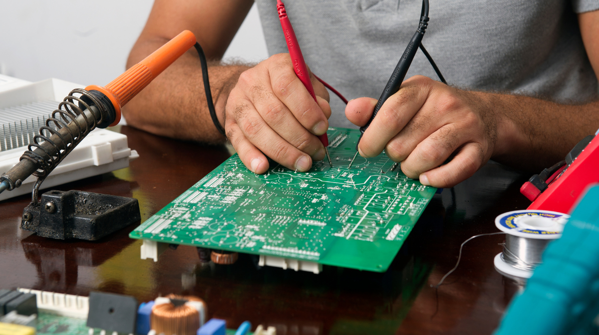 Person testing a printed circuit board with multimeter probes and nearby soldering tools.