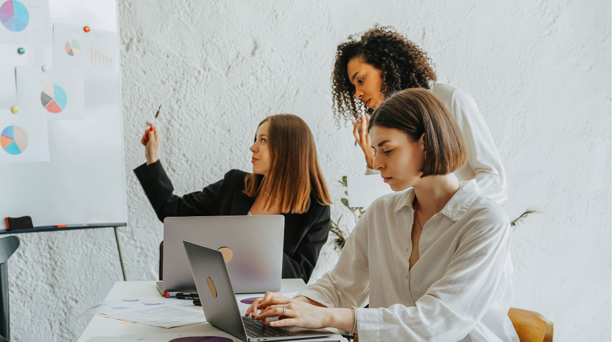 Three female colleagues collaborating at a table: one types on a laptop while another points to a whiteboard with charts and a third observes.