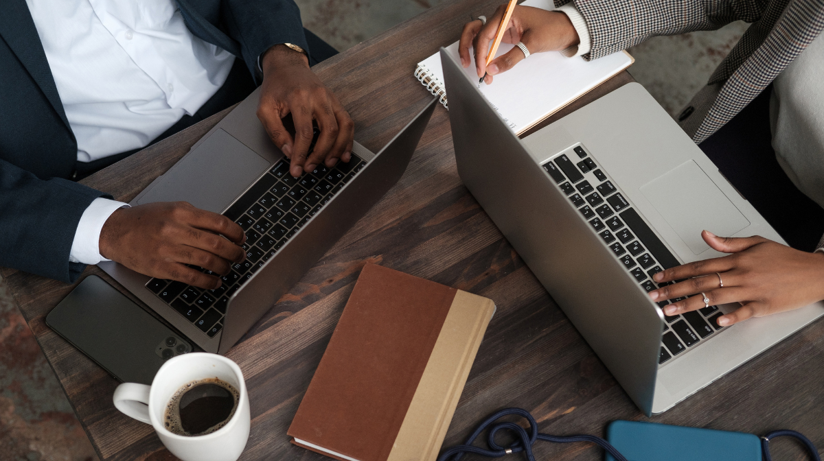 Two people working side-by-side at a table on laptops, one taking notes in a notebook.