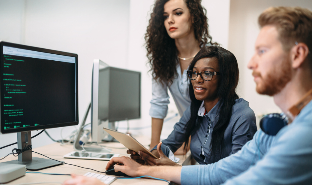 Three young professionals collaborating at a computer workstation, one typing while another holds a tablet and a third observes.
