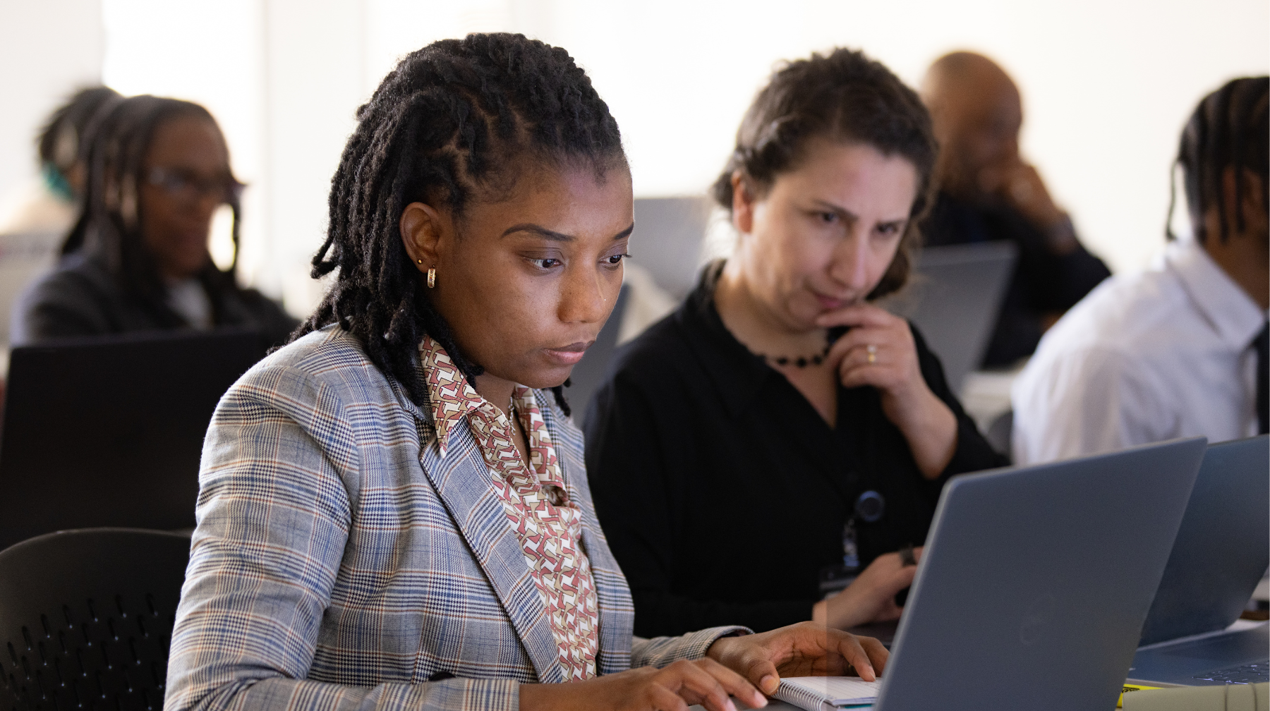 Two adult learners focused on laptops in a classroom, one typing while another looks on thoughtfully.