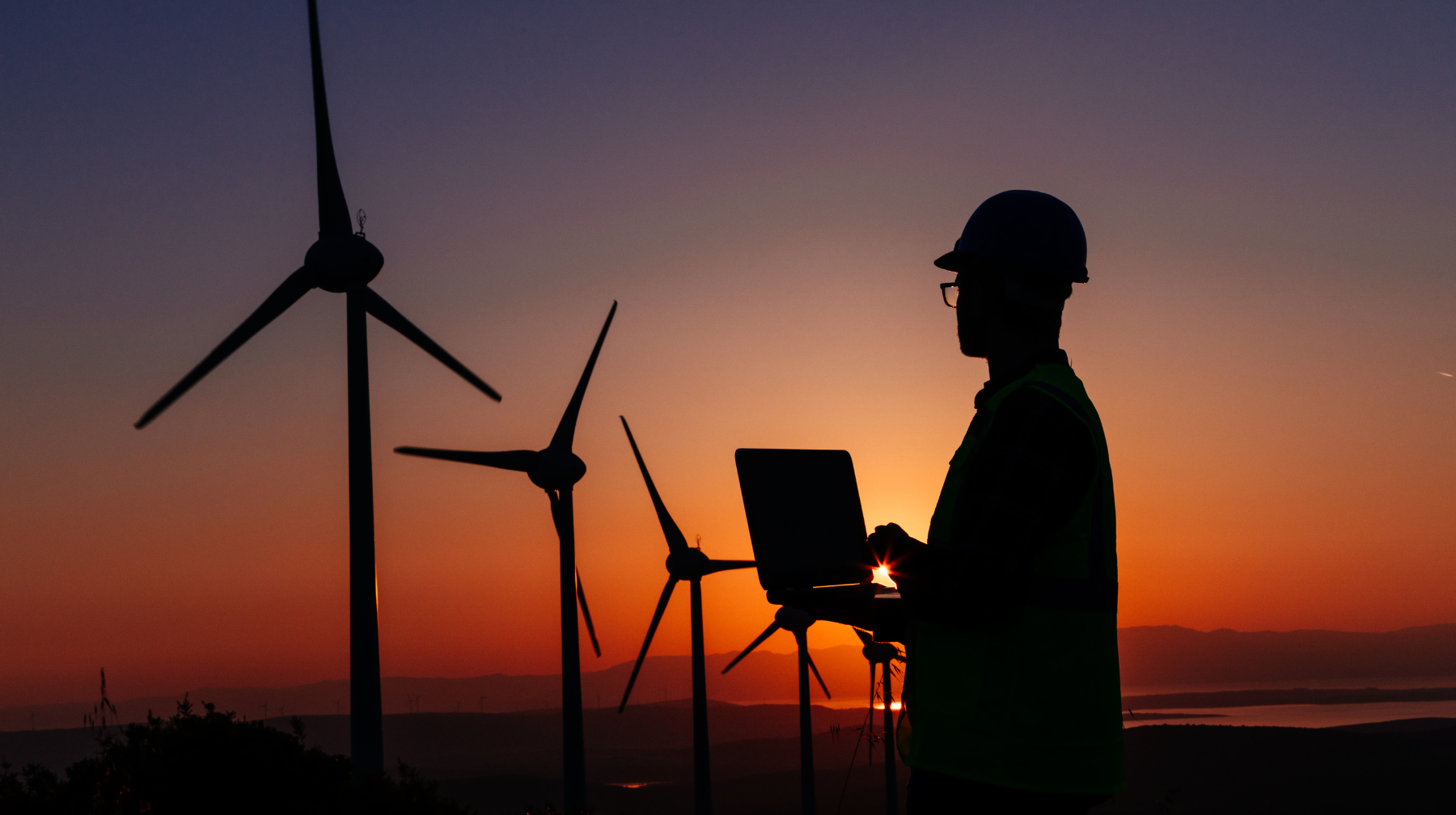 Silhouette of a wind farm technician holding a laptop while inspecting wind turbines at sunset.