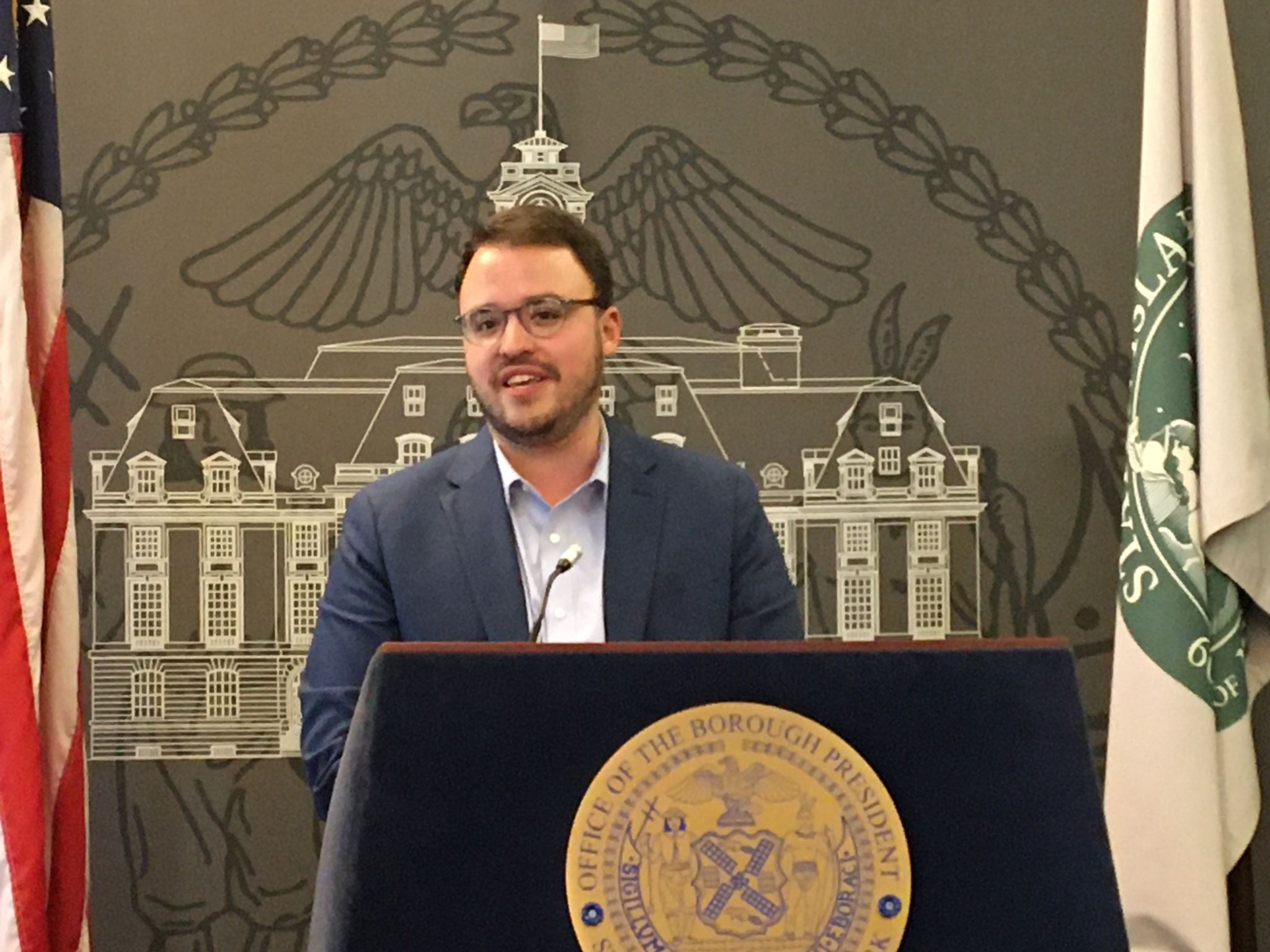 Male standing at a podium giving a speech, with a city seal on the podium and flags flanking a backdrop showing a building illustration.