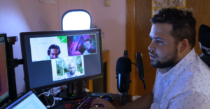 Man working at a home computer desk while participating in a video call shown on his monitor.