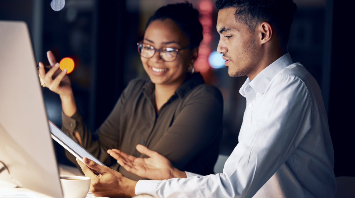 Two young adults collaborating as one holds a tablet in a dim office setting.