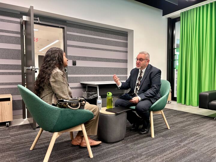 Two people sit in a small meeting area having a discussion, one holding a notebook and gesturing while the other listens.