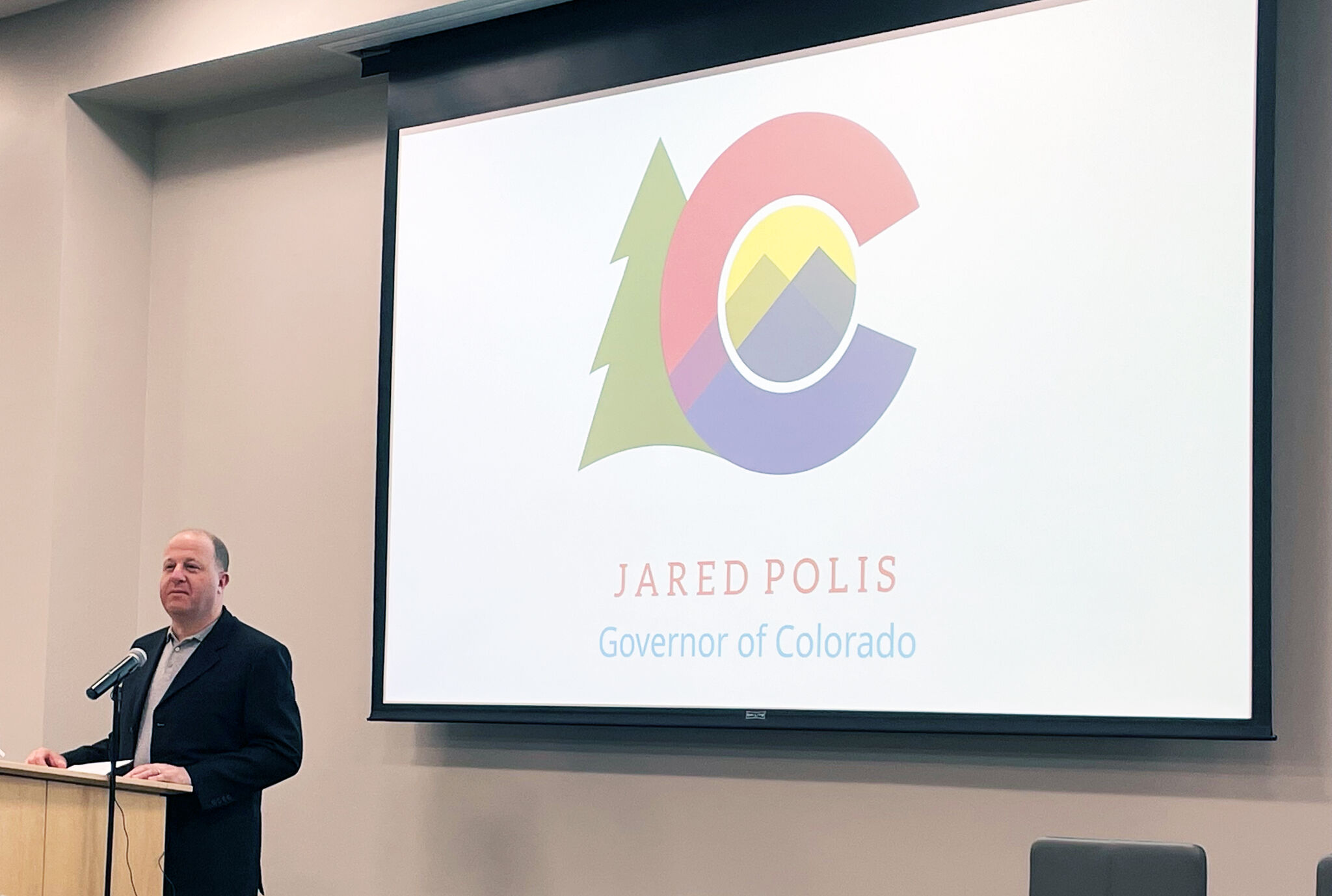 Person standing at a podium speaking beside a large screen displaying a logo and the text "Jared Polis Governor of Colorado."