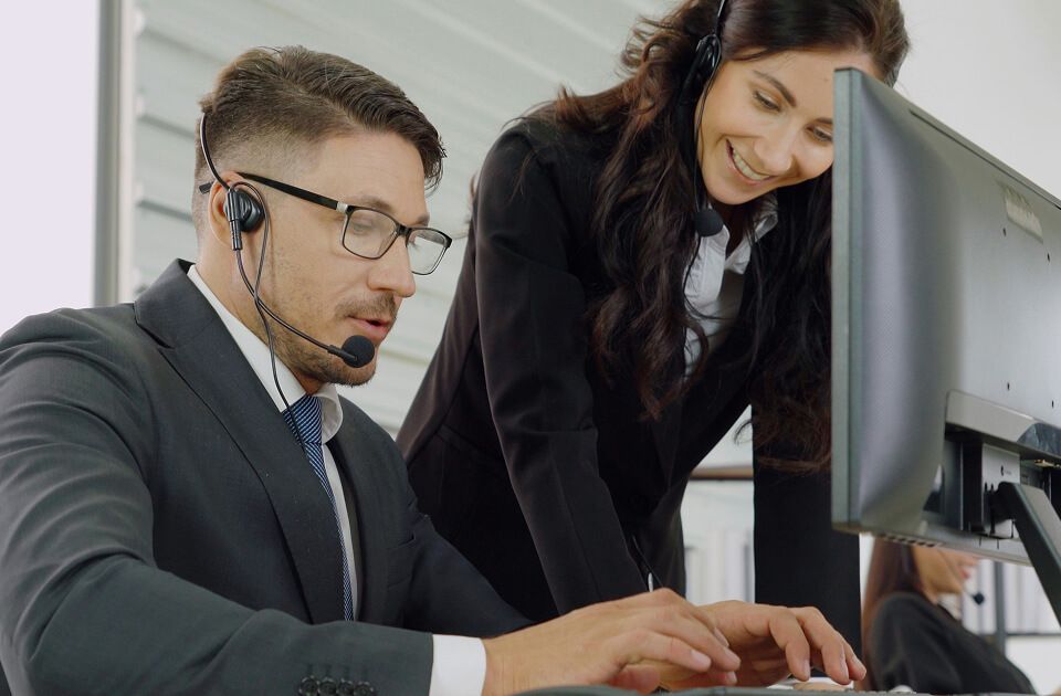A male and female wearing headsets collaborate in front of a computer monitor in a modern office setting.