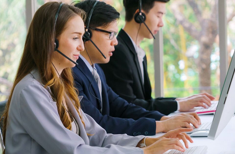 Three customer service agents wearing headsets typing at computers in an office with greenery visible through windows.