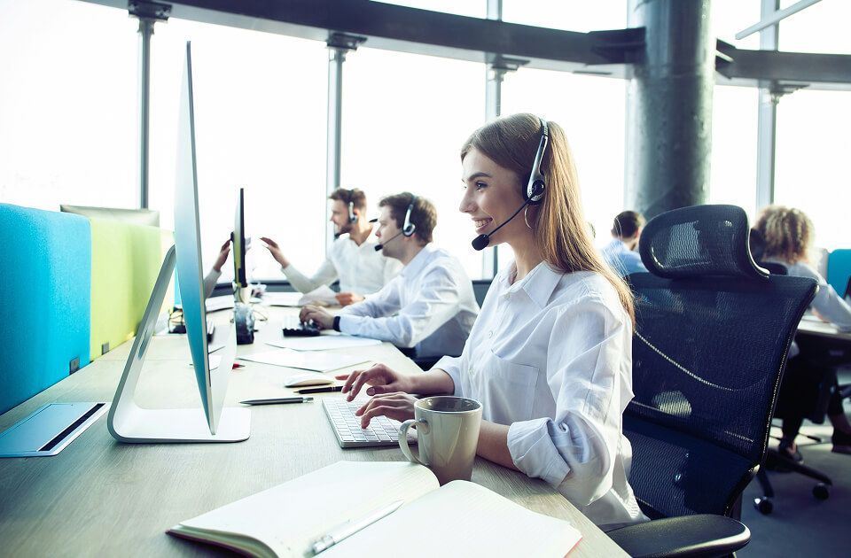 Smiling young professional wearing a headset, working at a desktop computer in a bright, modern call center.