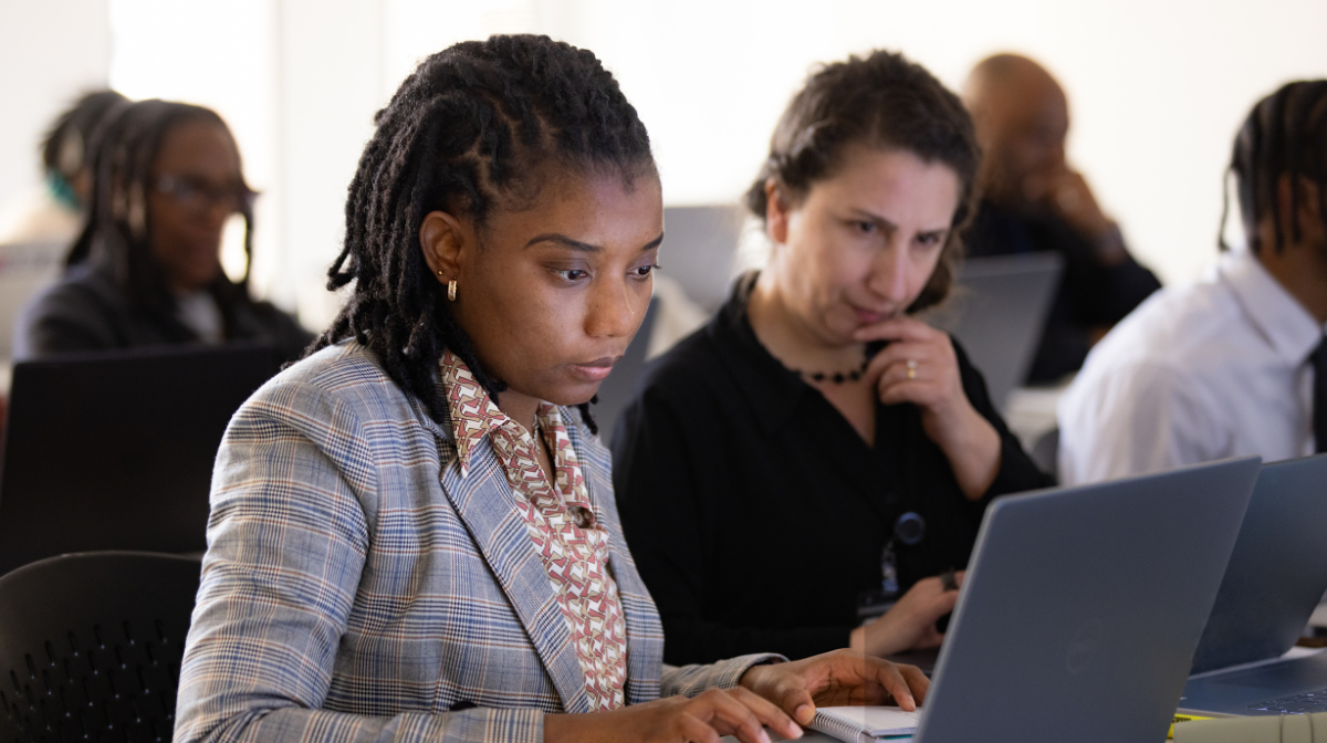 Two adult learners focused on laptops in a classroom, one typing while another looks on thoughtfully.