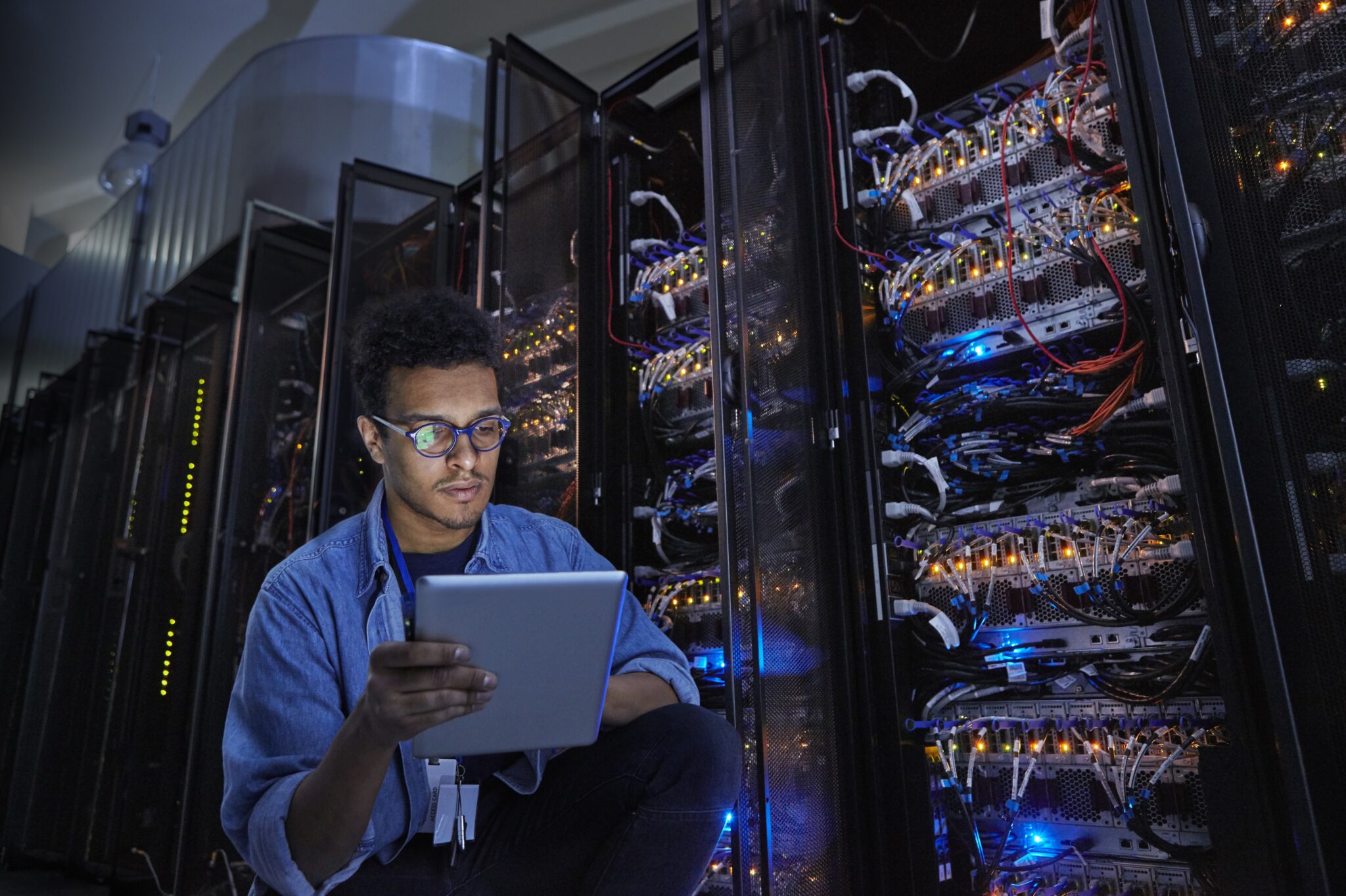 Technician kneeling in a server room, using a tablet while checking racks of network servers.