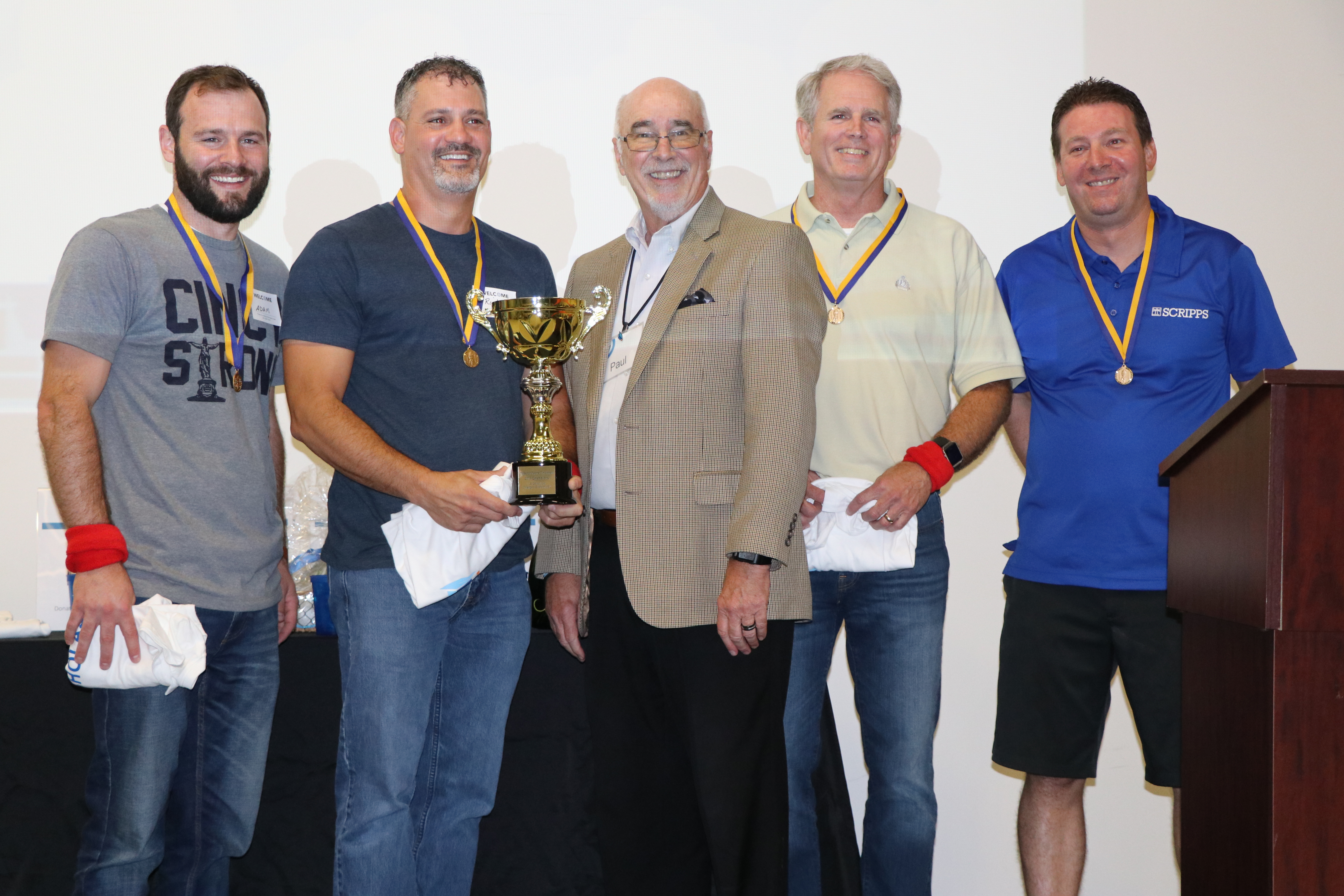 Five smiling men stand together receiving a trophy and wearing medals.