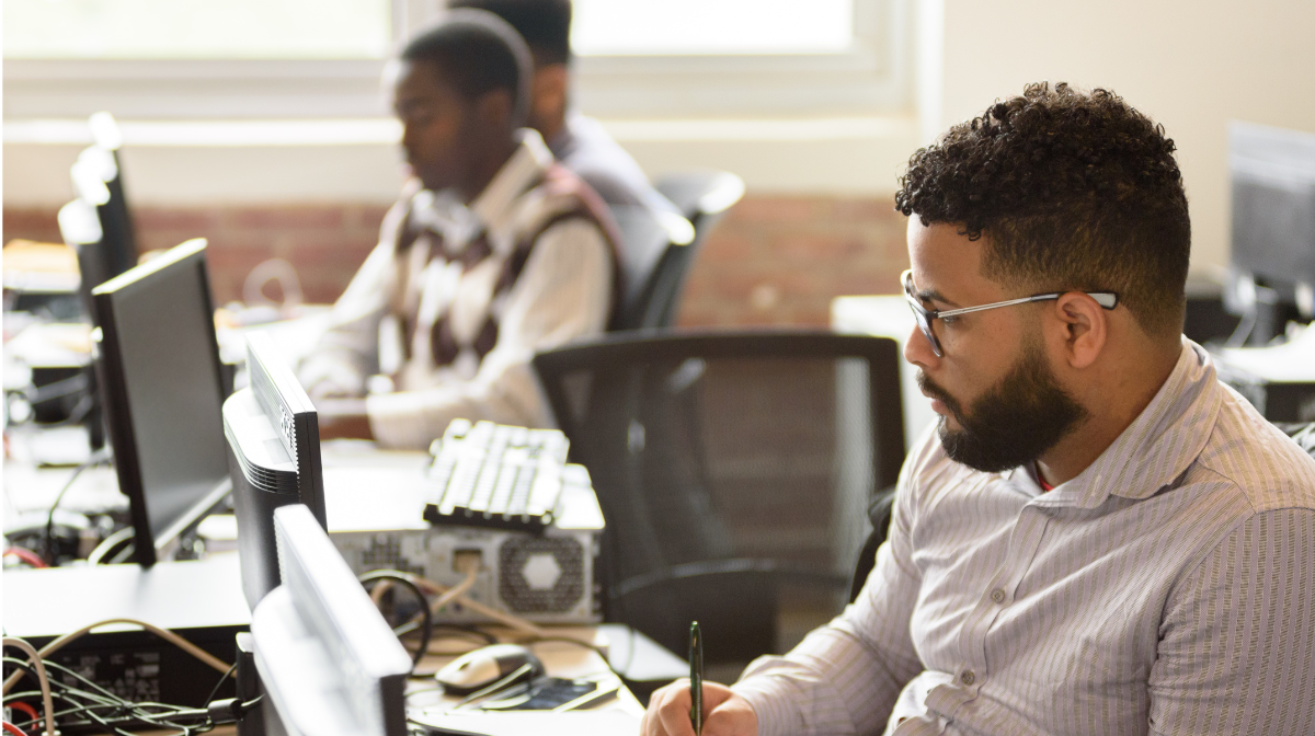 Man with beard and glasses writing notes at a dual-monitor desk in a bright office with brick walls, with colleagues working in the background.