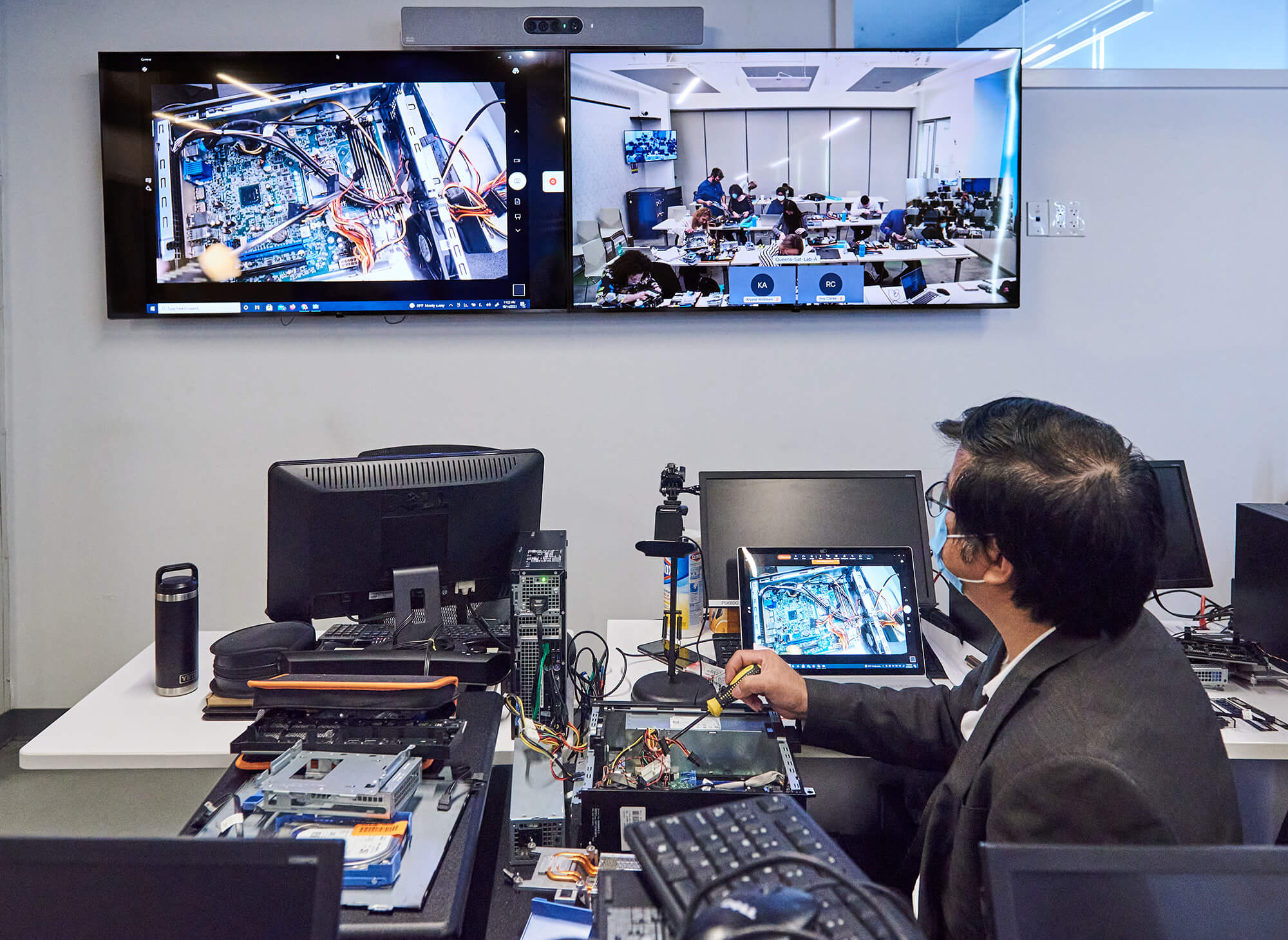 Instructor at a workstation demonstrating computer hardware troubleshooting on a livestream, with two large screens showing a close-up of internals and a classroom.