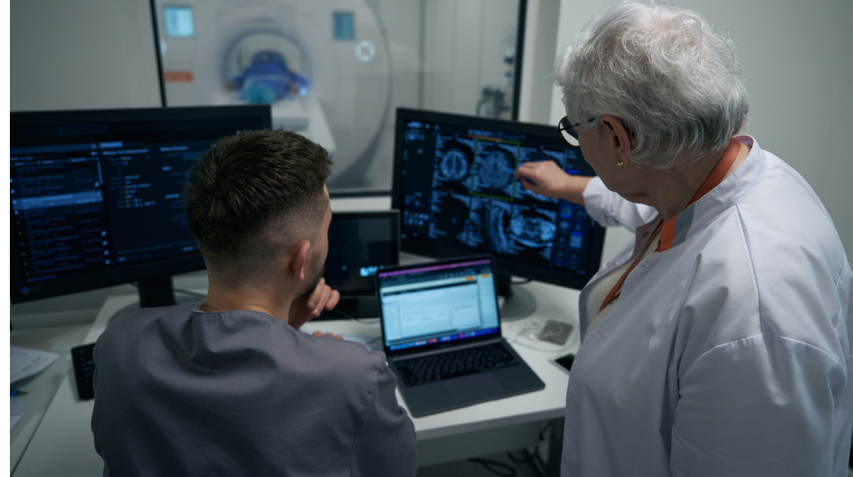 Two medical professionals review scans and patient data on multiple monitors beside an MRI scanner.
