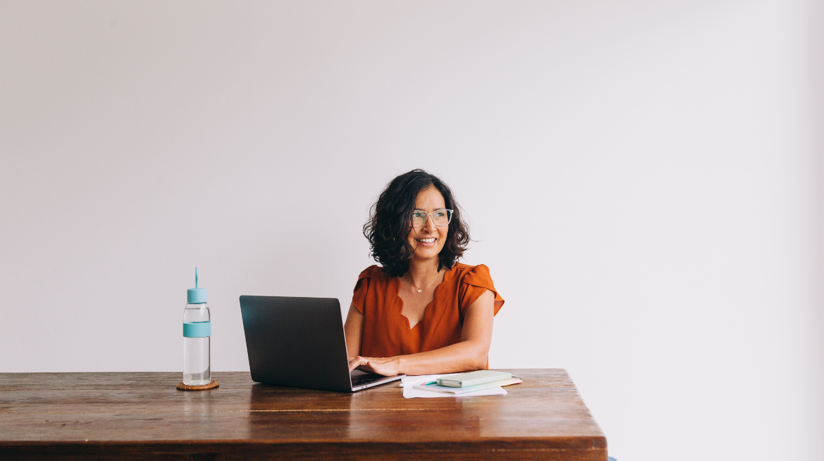 Woman sitting at a table working on a laptop and smiling, with a water bottle and notebooks nearby.