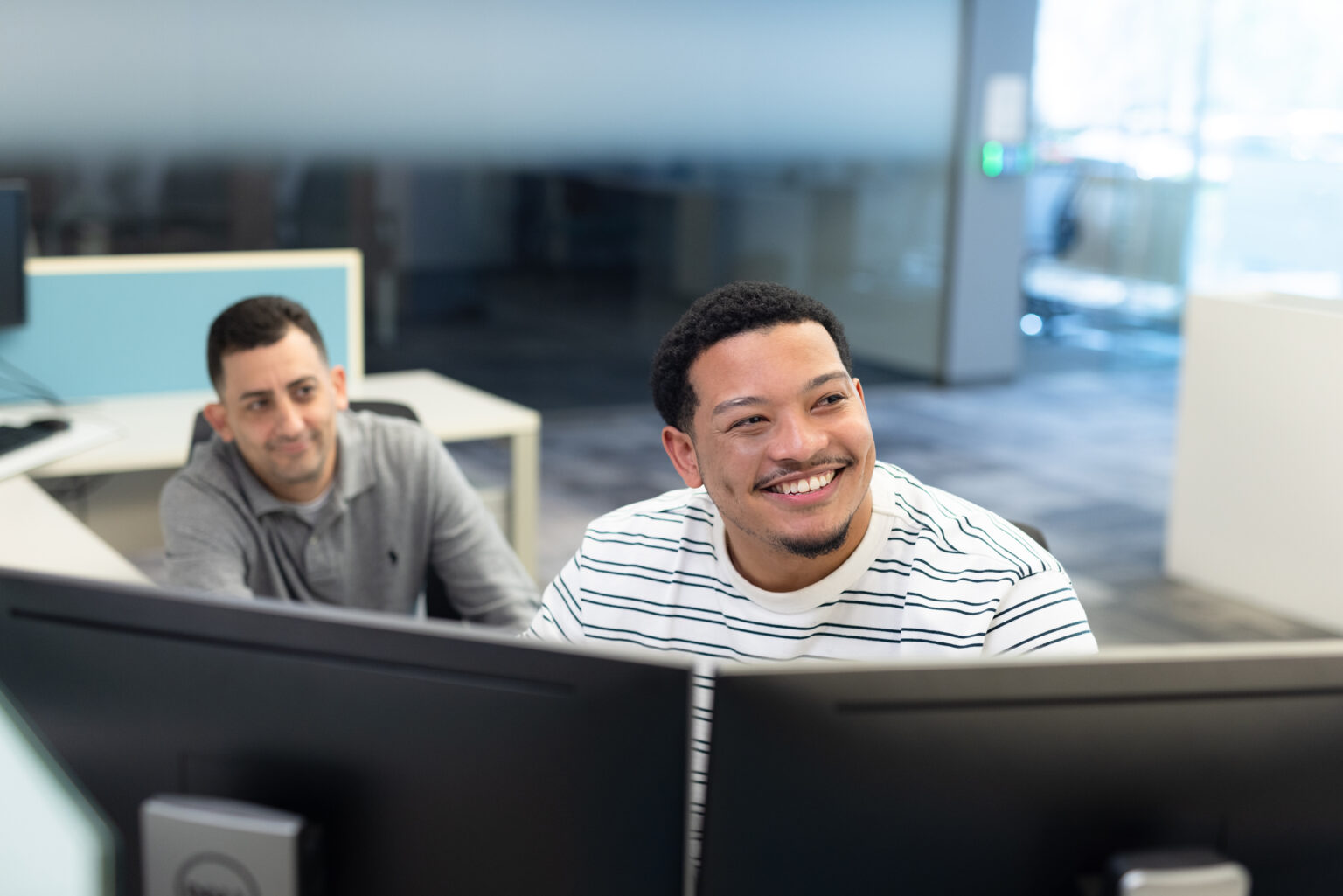 Two male colleagues smiling while working at desktop computers in a shared office space.