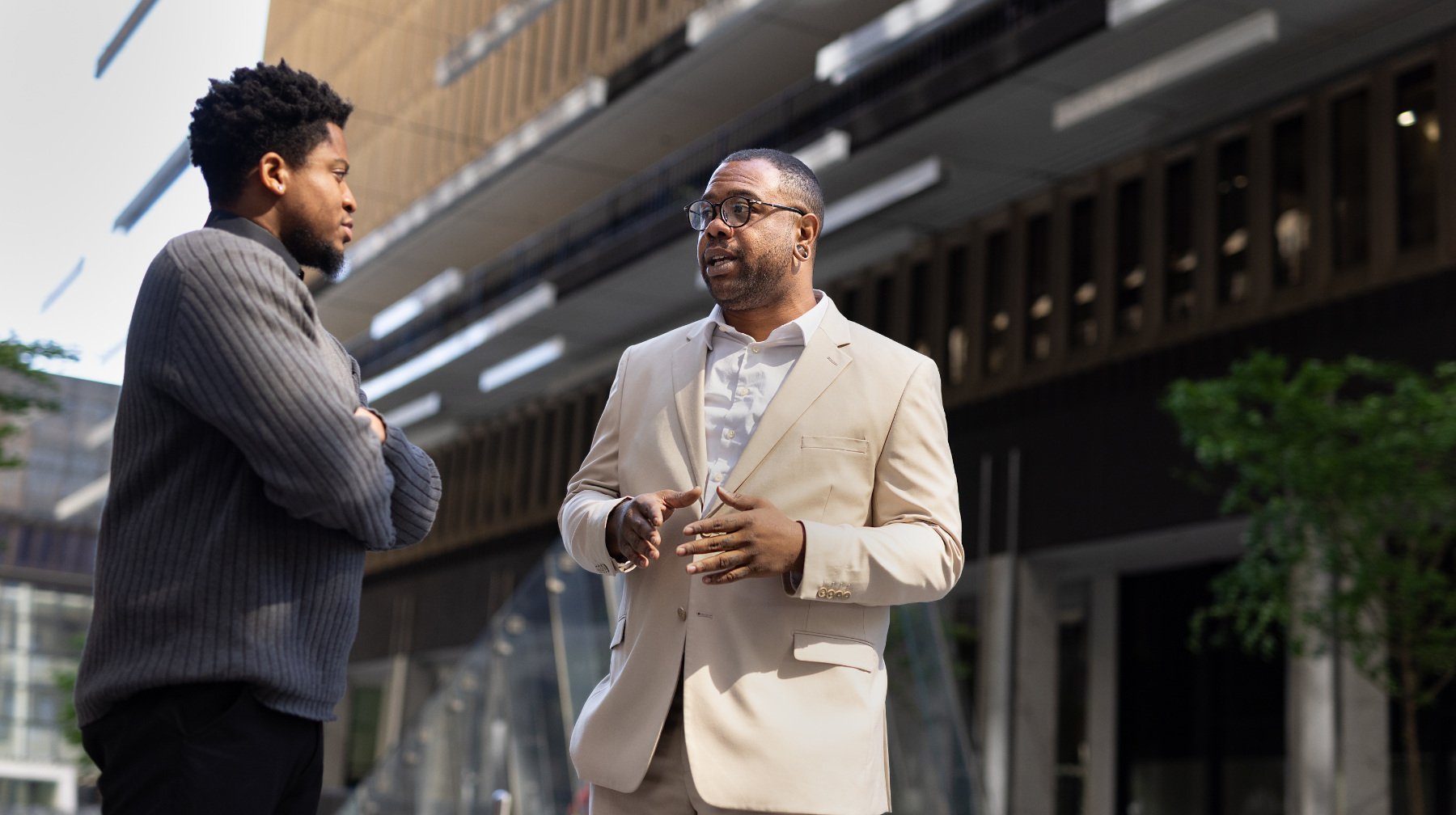 Two adults conversing outside an office building, one animatedly speaking while the other listens.