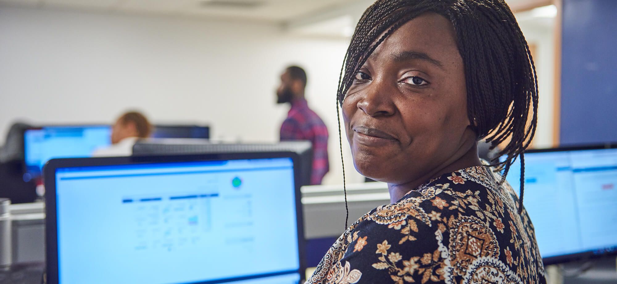 A student smiling at the camera while seated at a computer workstation in a classroom.