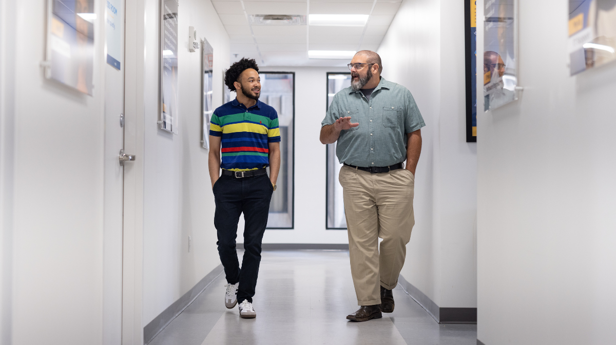 Young man in a striped polo shirt smiling and talking with an older man as they walk down a hallway.