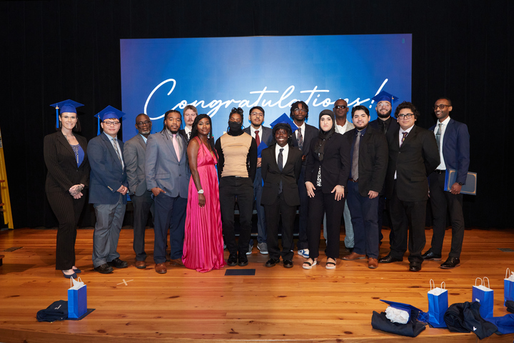 A diverse group of Per Scholas graduates and supporters standing onstage in front of a backdrop that says "Congratulations!".