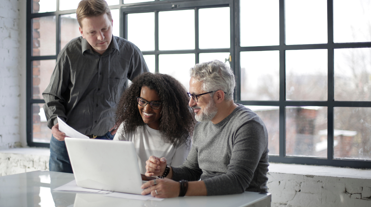 Three colleagues smiling and collaborating at a laptop by a large window.