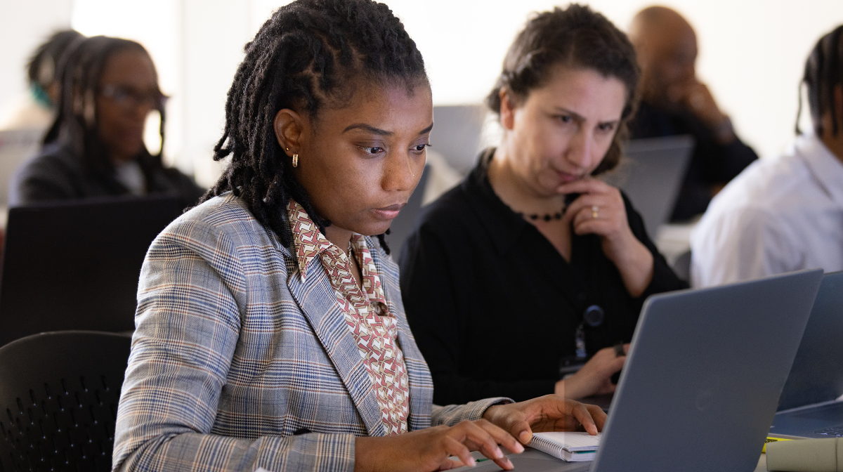 Two adult learners focused on laptops in a classroom, one typing while another looks on thoughtfully.
