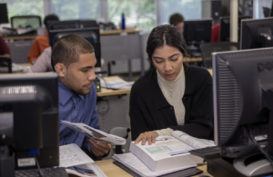 Two adult students at a computer workstation studying together, one pointing to a textbook as they discuss.