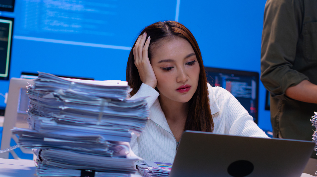 Young adult at a laptop looking frustrated while surrounded by tall stacks of paperwork in a tech workspace.