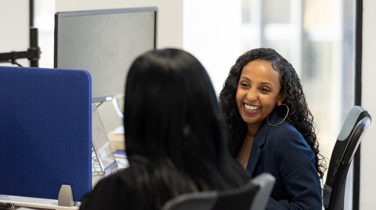 Two women smiling and talking at adjacent desks in an office setting.
