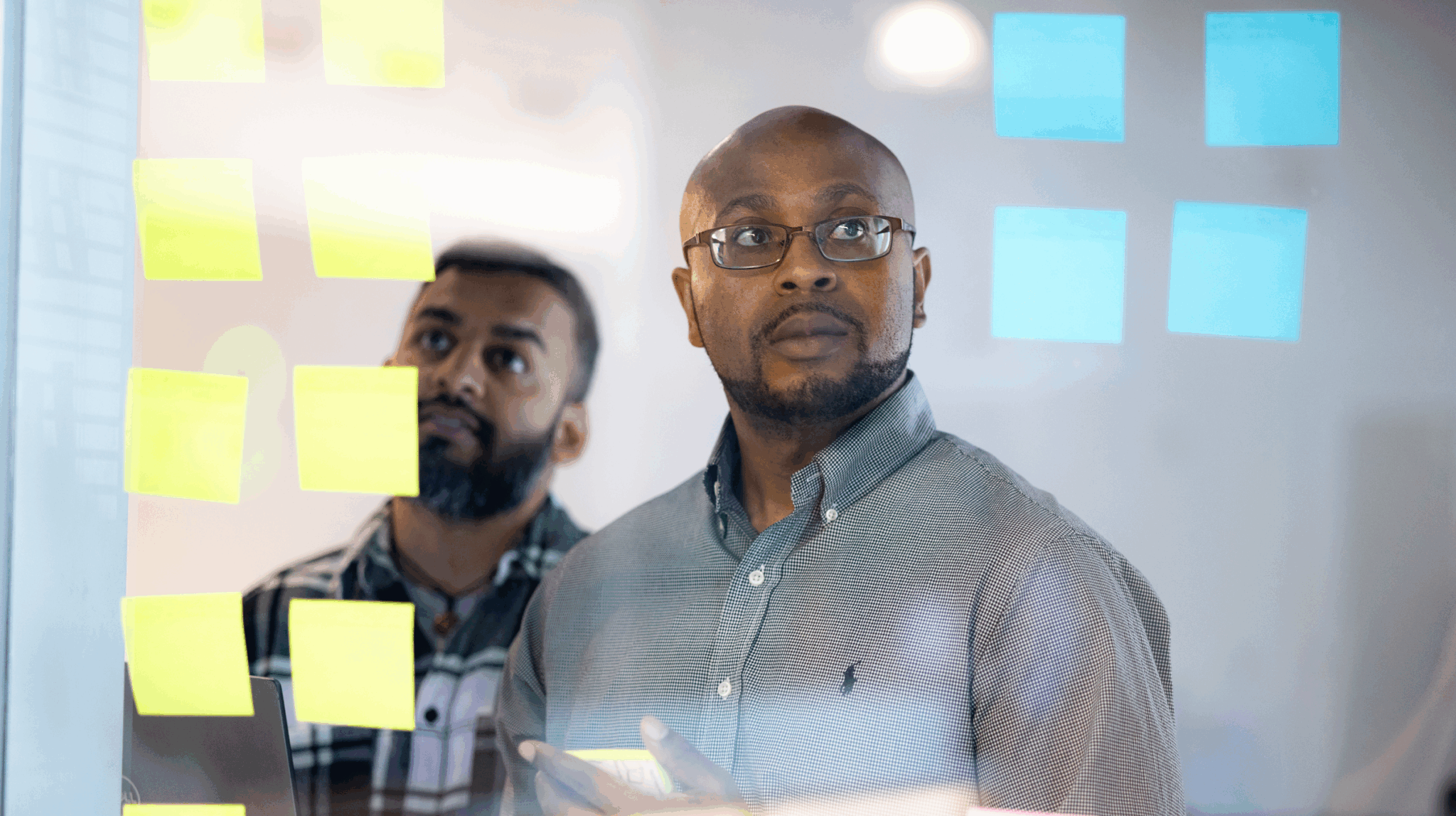 Two men standing behind a glass wall covered in sticky notes, looking thoughtfully at the notes.