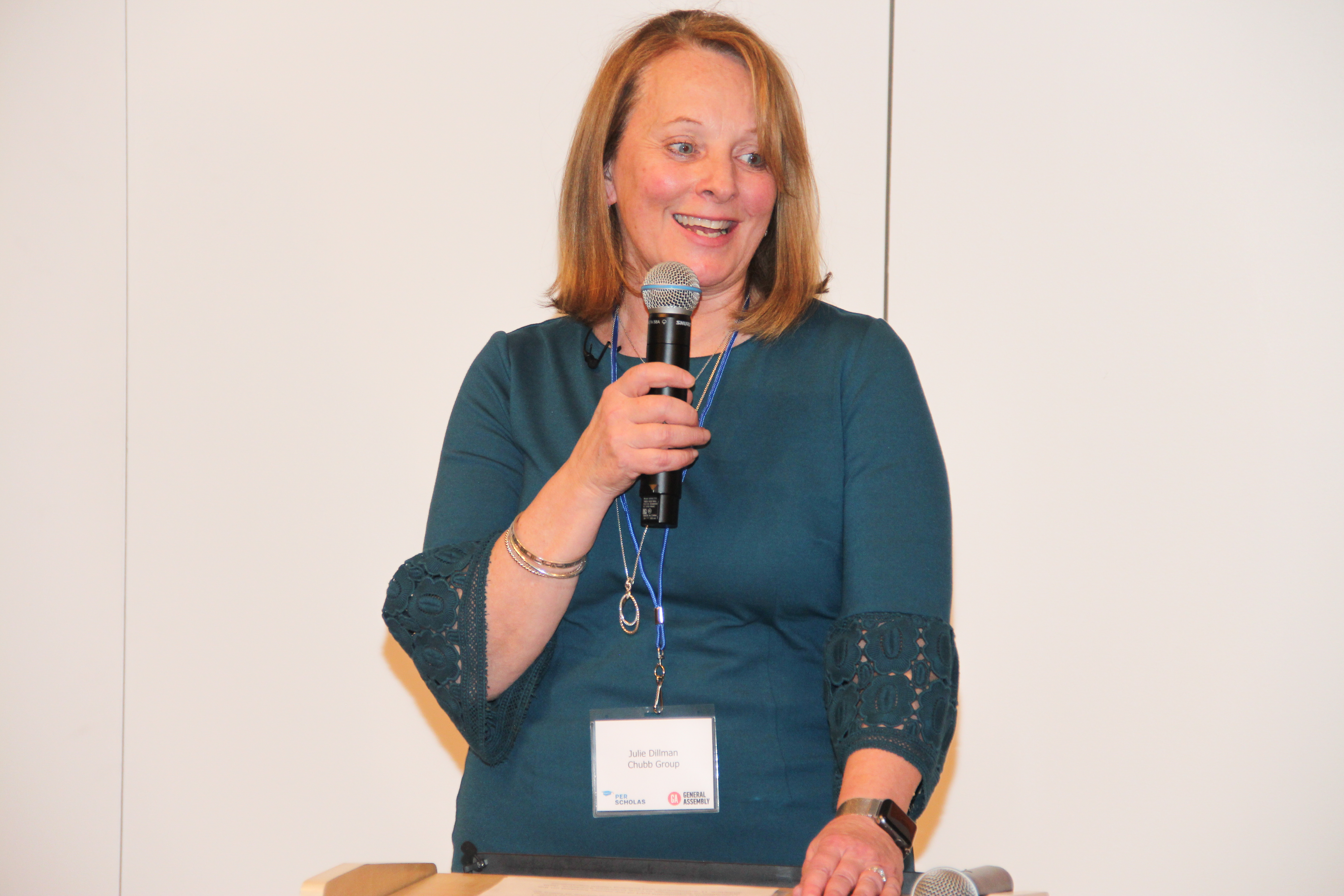 Woman speaking into a microphone at a podium, wearing a conference name badge.
