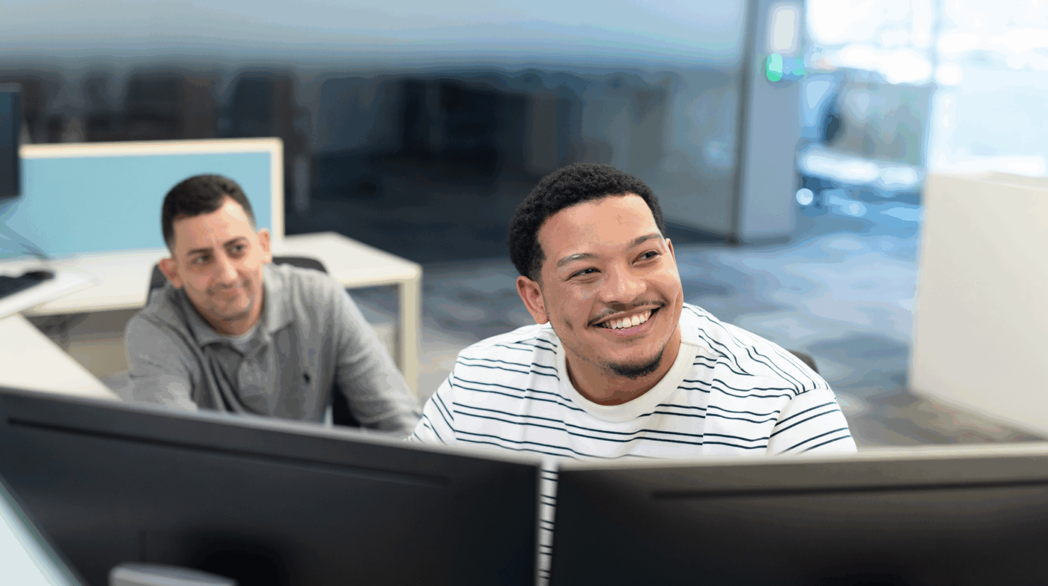 Two male colleagues smiling while working at desktop computers in a shared office space.