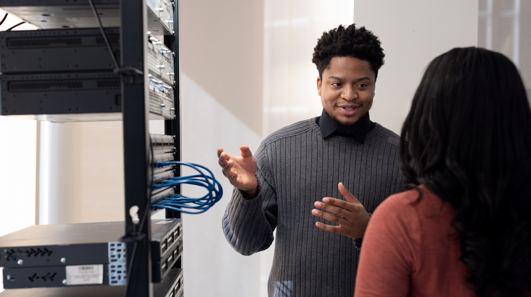 Two professionals talking beside a server rack, with one person explaining and gesturing to the other.