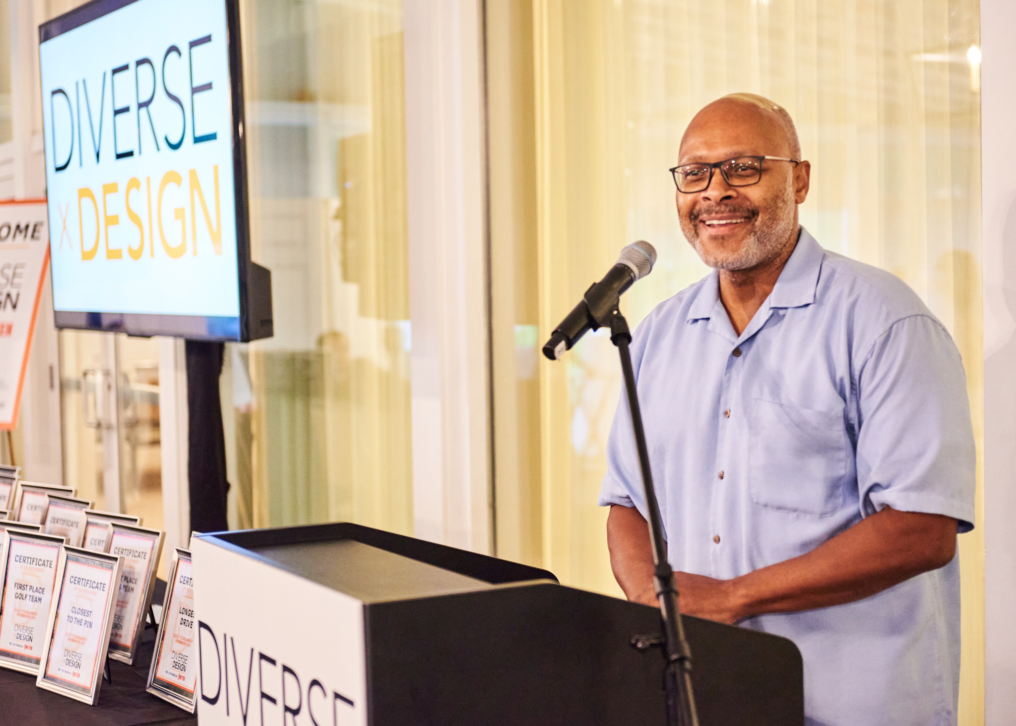 Man speaking at a podium with a microphone and a "Diverse x Design" sign next to him.