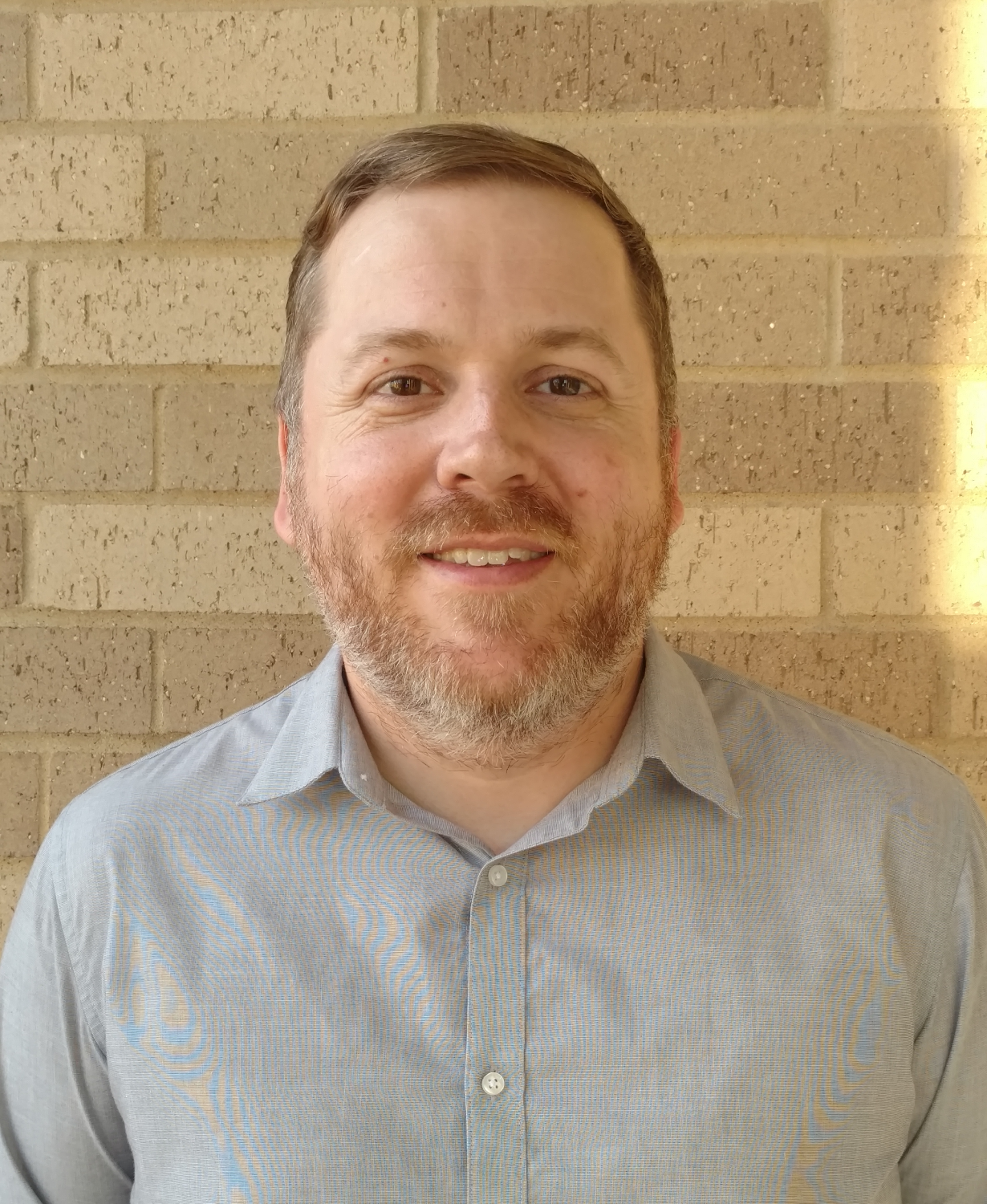 Smiling adult man posed against a brick wall, shown from chest up.
