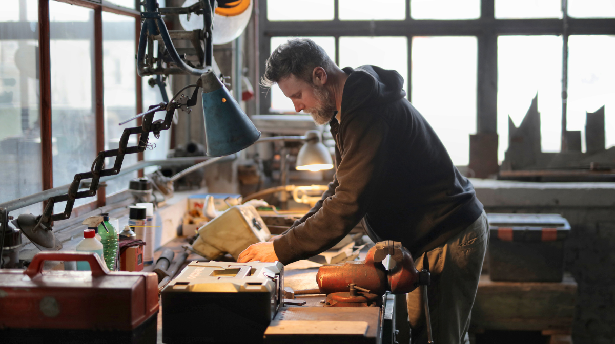 Adult man leaning over a workshop bench working with tools under task lamps.
