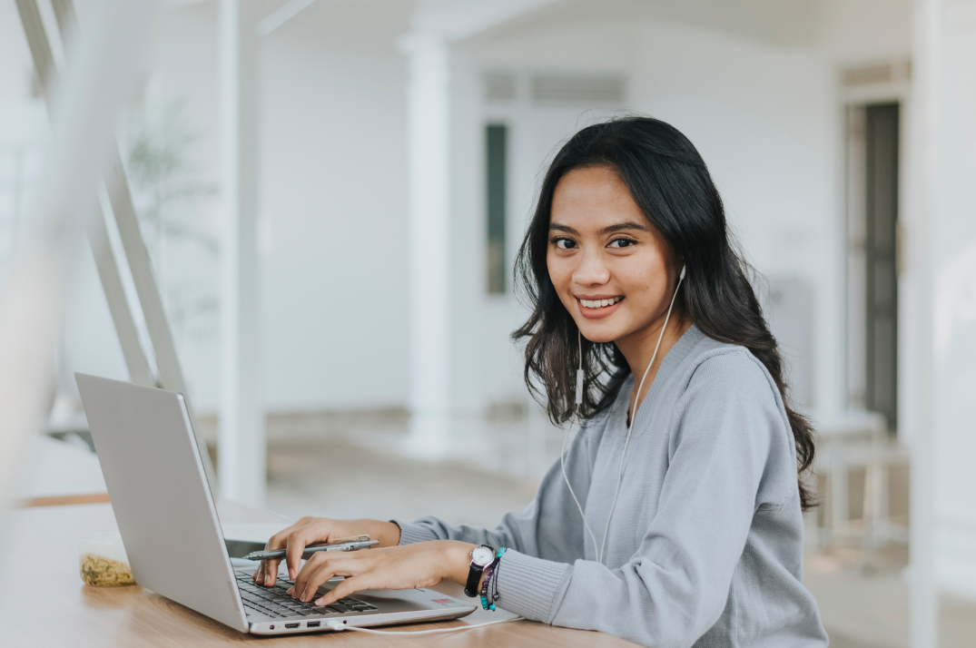 Young woman smiling while typing on a laptop with earbuds in, seated at a table.