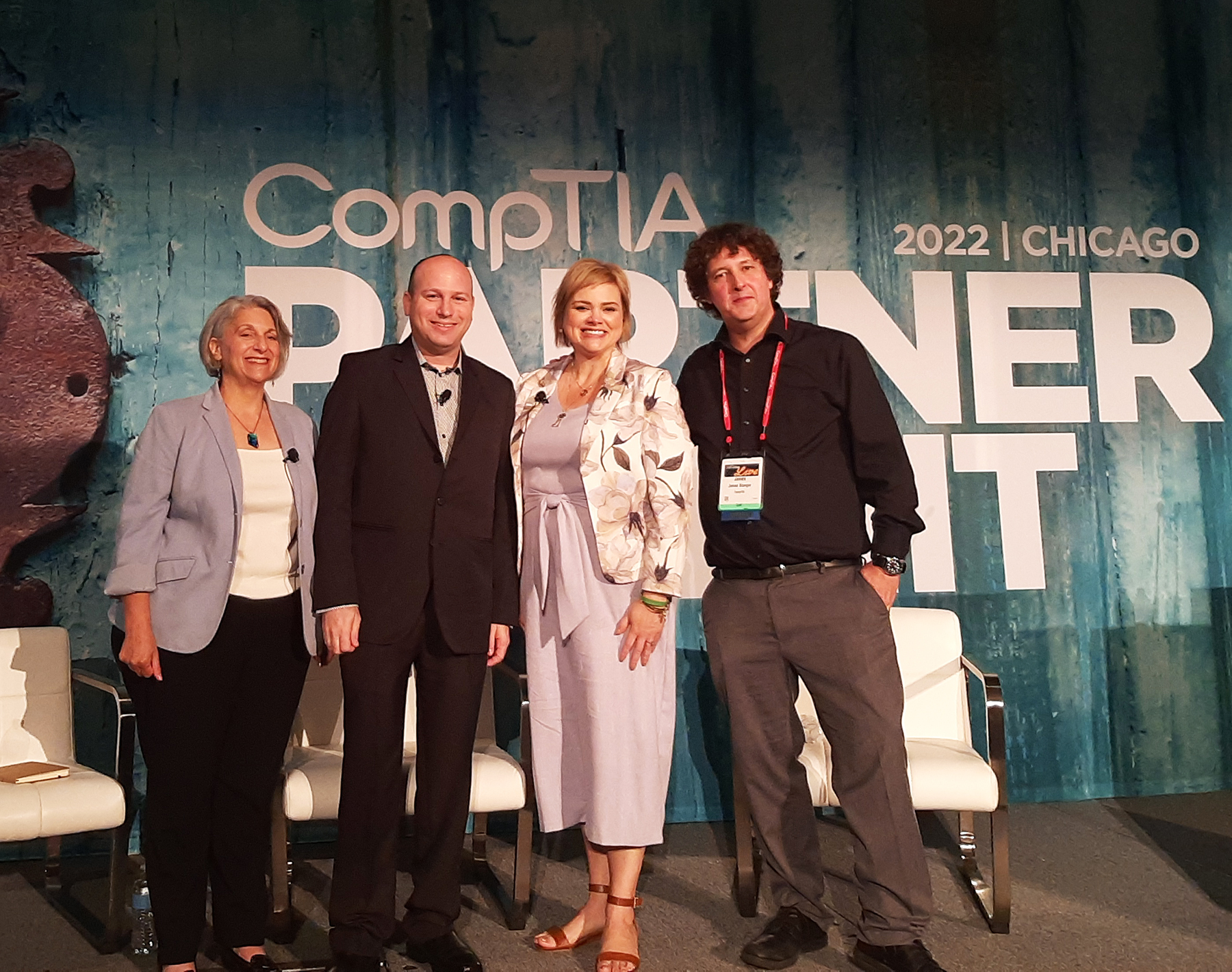 Four smiling conference speakers stand on stage in front of a backdrop reading "CompTIA Partner 2022 | Chicago."