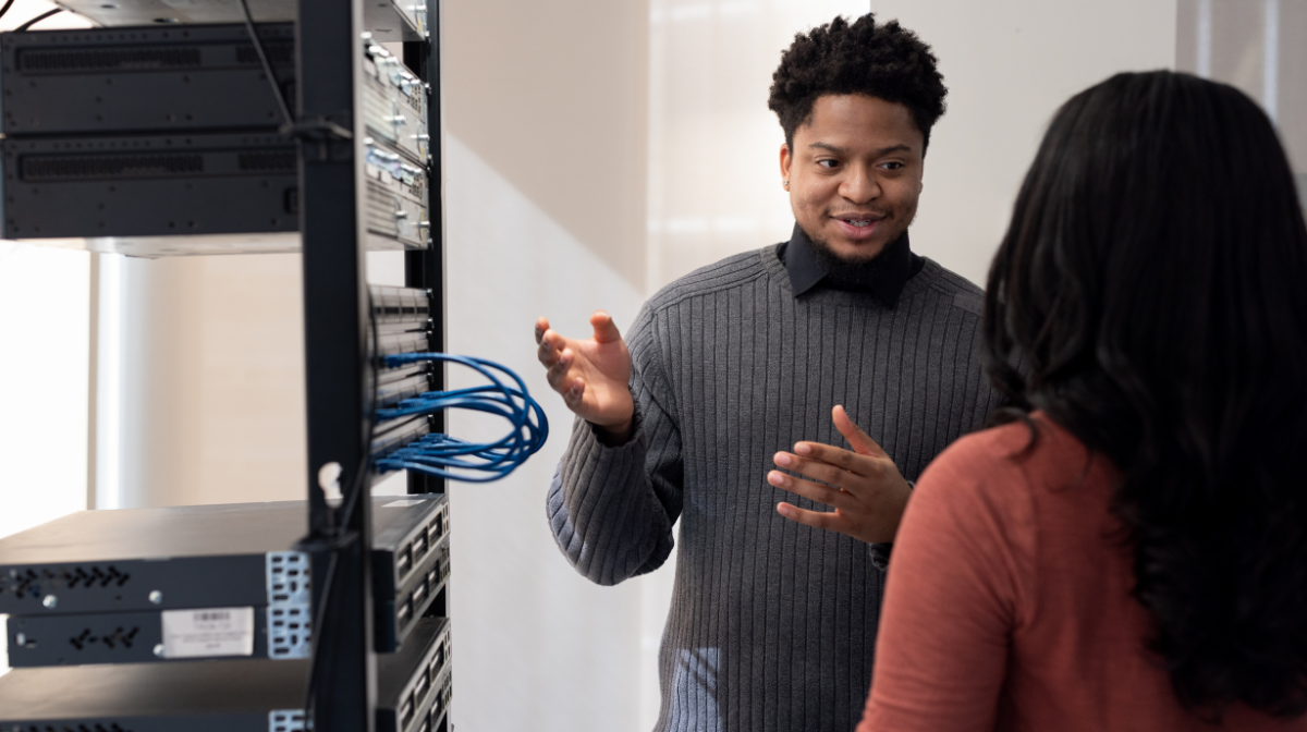 Two professionals talking beside a server rack, with one person explaining and gesturing to the other.