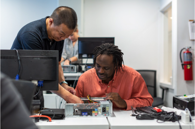 Two adult learners work together on an open desktop computer on a classroom table while another student and monitors are in the background.