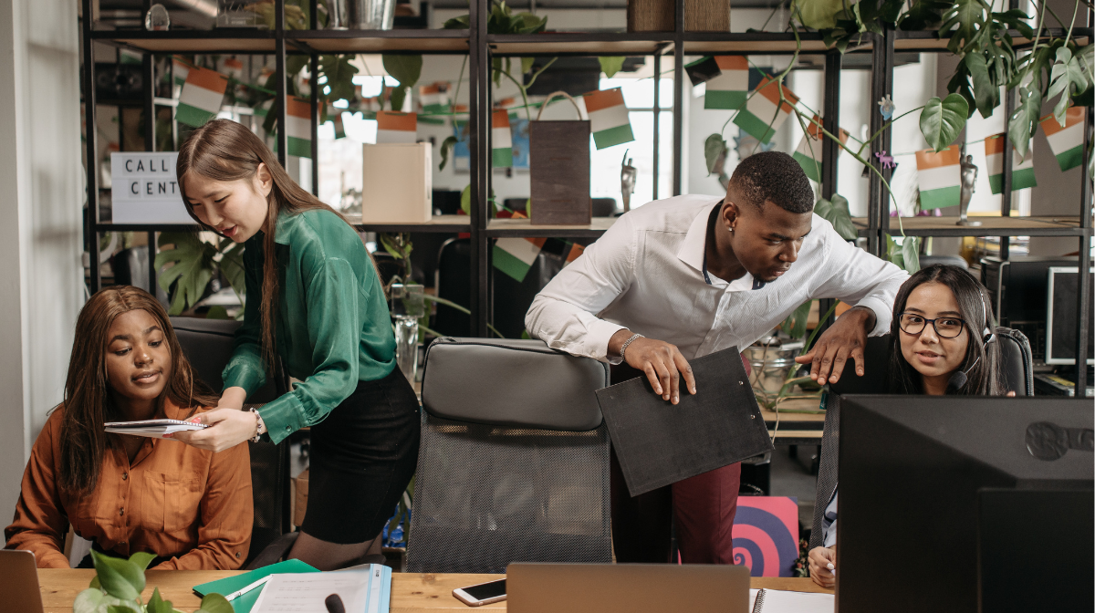 Four young professionals collaborating at a shared workspace, two standing and guiding coworkers seated at computers.