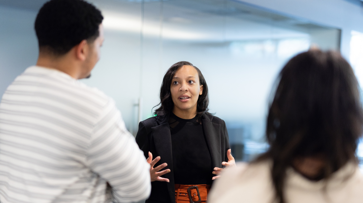 Woman speaking and gesturing to two colleagues in a workplace conversation.