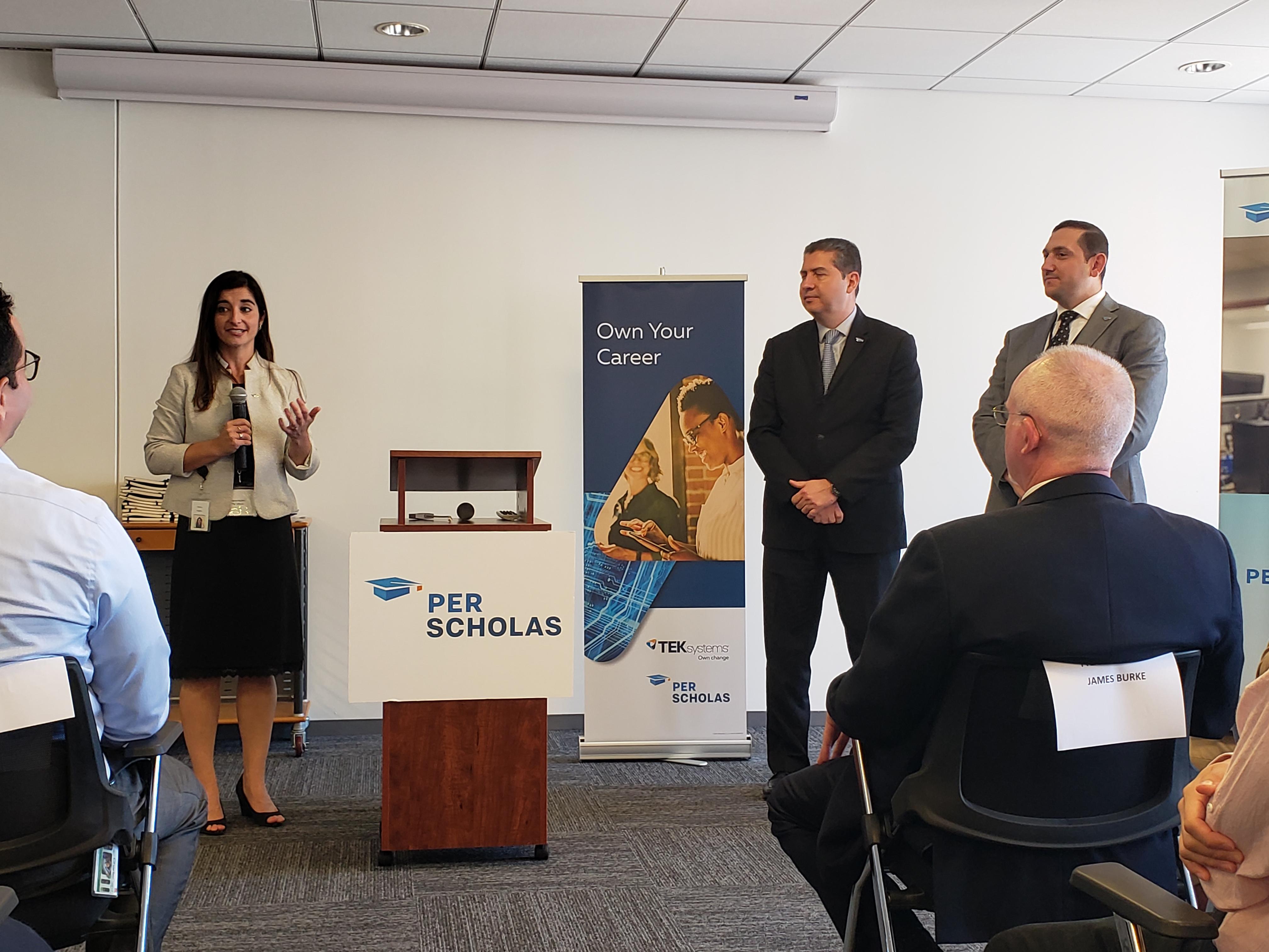 Woman speaking at a Per Scholas podium during a small panel event with two men standing nearby and seated attendees.