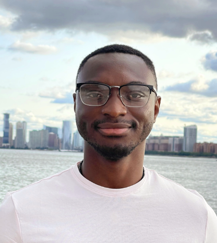Head-and-shoulders portrait of a smiling young man standing outdoors with a city skyline and water behind him.
