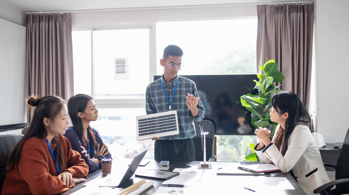 Young professional presenting a small solar panel and model wind turbine to colleagues around a conference table.