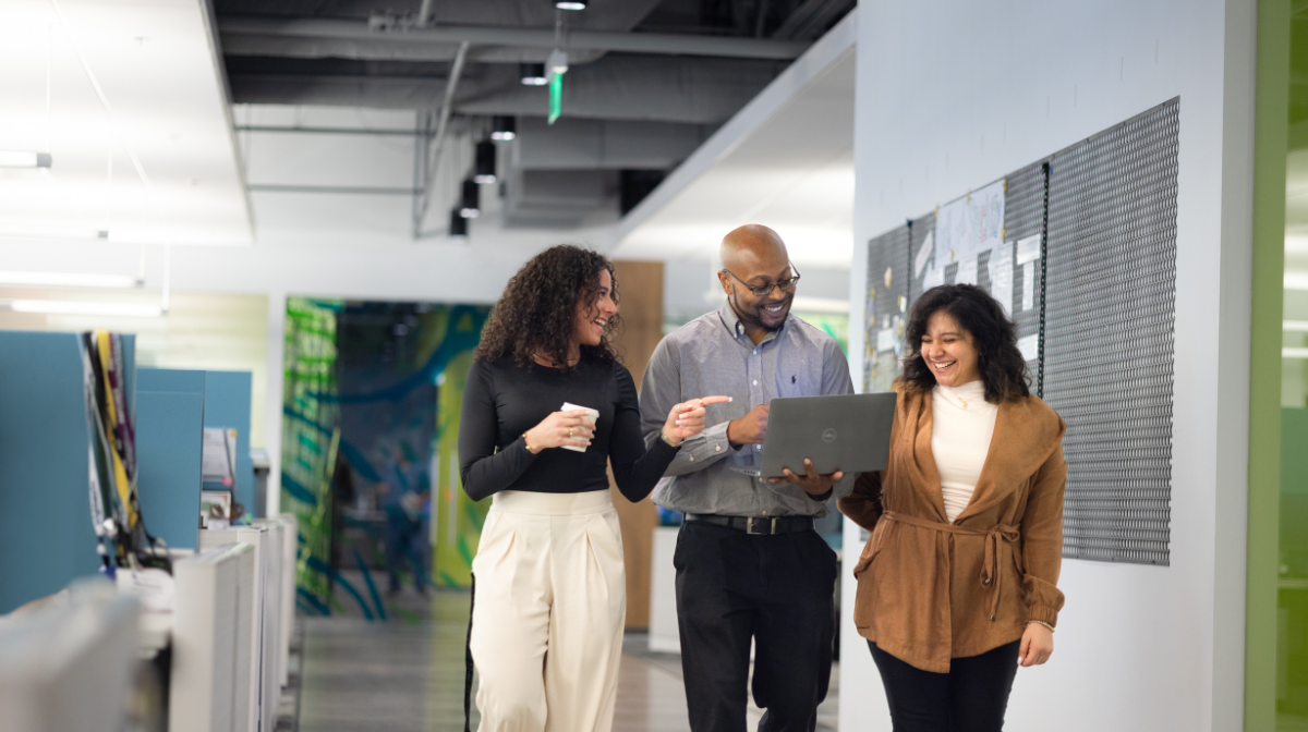 Three colleagues walking down an office hallway, smiling and discussing something on a laptop one person is holding.