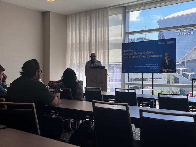 Presenter speaking at a conference podium with slide titled "Building a Military Ready Culture vs. Military Friendly Culture."